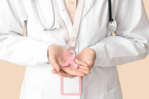 Female doctor with pink ribbon on beige background, closeup. Breast cancer awareness concept