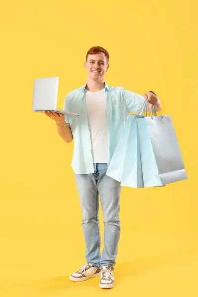 快乐的红头发年轻人 带着笔记本电脑和黄色背景的购物袋 黑色星期五发售 — 图库照片
