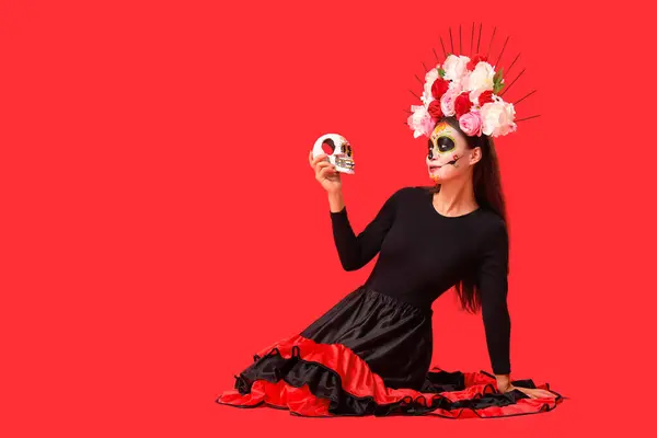 stock image Young woman with painted skull sitting on red background. Mexico's Day of the Dead (El Dia de Muertos) celebration