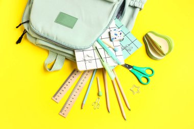 Sarı arka planda farklı kırtasiye malzemelerinin bulunduğu şık bir okul çantası.