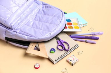 Bej arka planda farklı kırtasiye malzemesi içeren şık bir okul çantası.