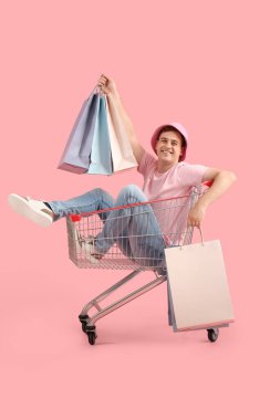 Çantalı mutlu genç adam pembe arka planda alışveriş arabasında oturuyor. Kara Cuma indirimi