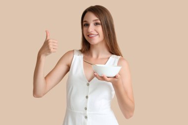 Tavuk çorbası içen genç bir kadın bej arka planda baş parmağını kaldırıyor.