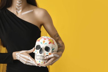 Cadılar Bayramı partisi için ölü gelin gibi giyinmiş genç bir kadın. Sarı arka planda insan kafatası resmi var.