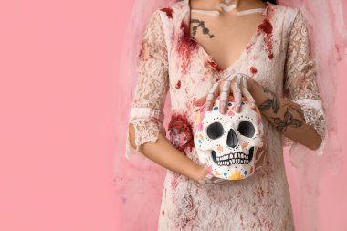 Cadılar Bayramı partisi için ölü gelin gibi giyinmiş genç bir kadın. Pembe arka planda insan kafatası resmi.