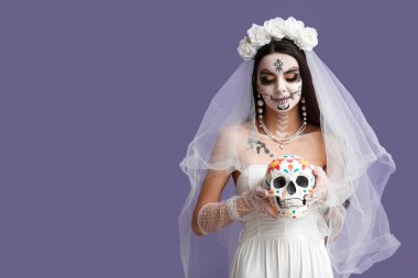 Cadılar Bayramı partisi için ölü gelin gibi giyinmiş genç bir kadın ve mor arka planda insan kafatası resmi.
