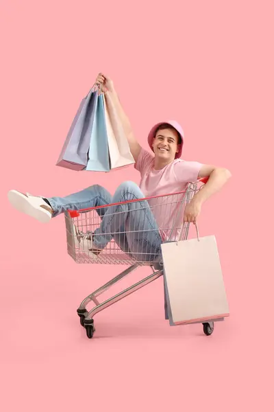 快乐的年轻人 提着袋子坐在粉色背景的购物车里 黑色星期五发售 — 图库照片