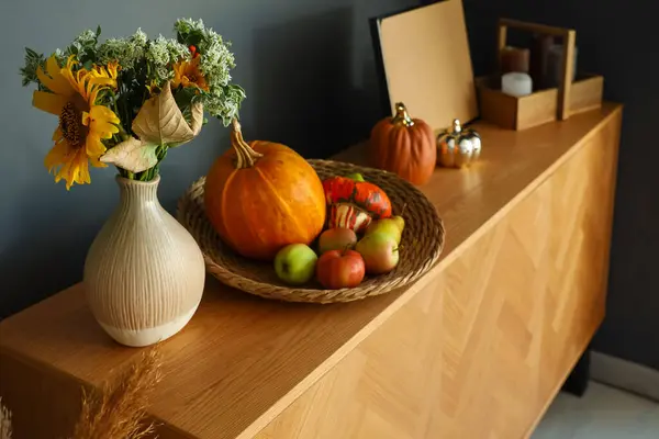 Vase with autumn flowers, pumpkins and apples on chest of drawers in room, closeup