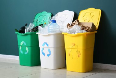 Geri dönüşüm sembollü çöp kutuları ve yeşil duvarın yanındaki farklı çöpler.