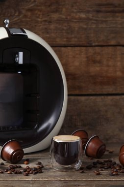 Bir bardak nefis espresso, kapsül kahve makinesi, kapsüller ve fasulyeler ahşap masada.