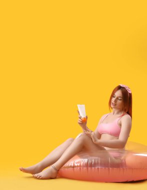 Şişirilebilir yüzüklü genç bir kadın sarı arka plana güneş kremi sürüyor.