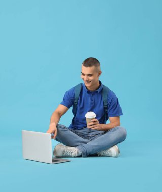 Mavi arka planda oturan ve dizüstü bilgisayar kullanan mutlu erkek öğrenci.