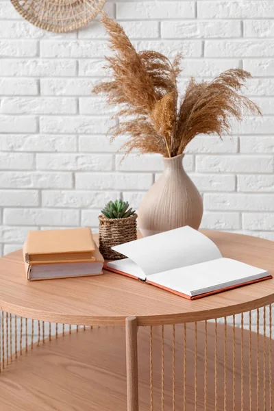 Light wooden coffee table with vase of dried pampas grass and books near white brick wall
