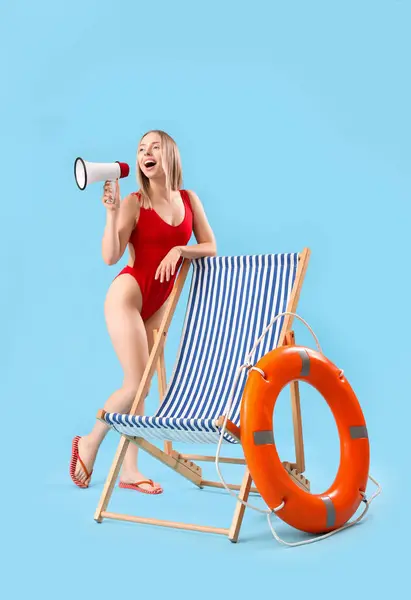 Beautiful woman with megaphone, deck chair and ring buoy on blue background