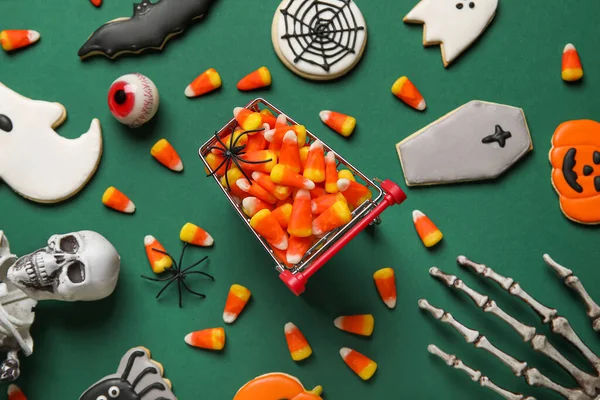 Shopping cart with tasty candy corns, cookies for Halloween and skeleton on green background