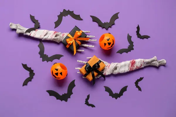 Composition with skeleton hands, gift boxes, pumpkins and paper bats for Halloween celebration on purple background
