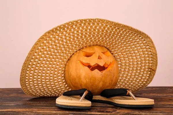 Carved pumpkin for Halloween with stylish hat and flip flops on table near beige wall