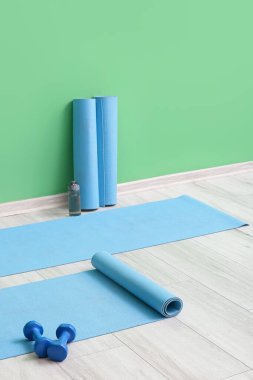 Spor salonundaki yeşil duvarın yanında dambılı ve şişeli yoga minderleri.