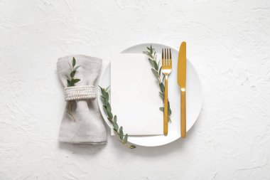 Boş davetiye kartı, çatal bıçak takımı ve beyaz masa üzerinde okaliptüs dallarıyla temiz tabak.