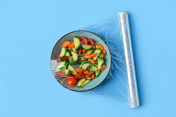 Bowl with salad wrapped with stretch wrap on blue background