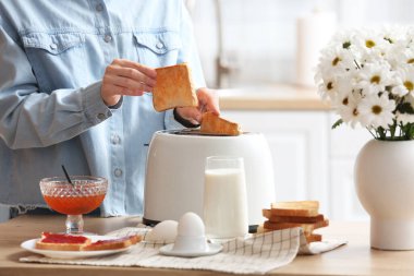 Çağdaş ekmek kızartma makinesinden çıtır ekmek dilimleri alan bir kadın mutfakta kahvaltıyla masada oturuyor.