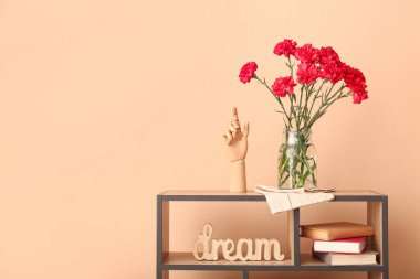 Bej duvarın yanındaki rafta kırmızı karanfilli vazo.