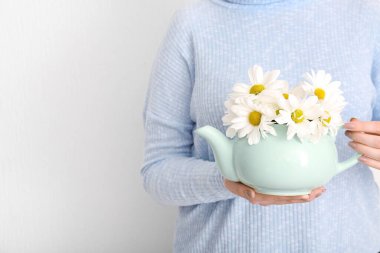 Beyaz duvarın yanında çaydanlık dolusu papatya çayı ve çiçeklerle güzel genç bir kadın.