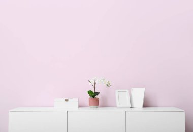 Orkide çiçekli beyaz çekmece ve odada pembe duvarın yanında boş resimler.