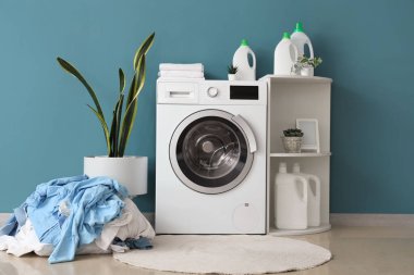 Çamaşır makinesi, raf ünitesi, ev bitkisi ve mavi duvarın yanındaki kirli çamaşır yığını.