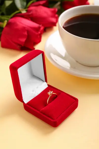 Box with engagement ring, cup of coffee and roses on beige background, closeup
