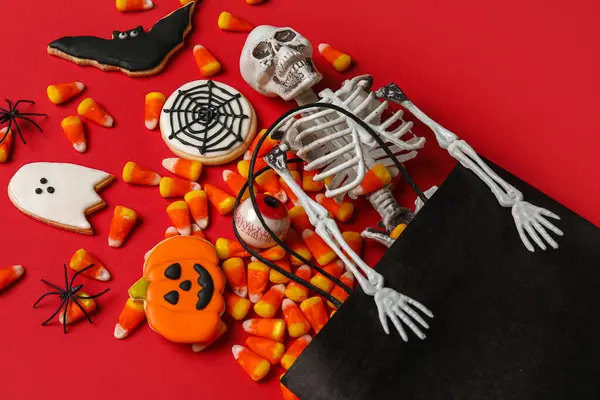 Shopping cart with tasty candy corns, skeleton and cookies for Halloween on red background