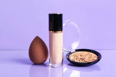 Mor zemin üzerinde sünger ve yüz pudrası olan bir şişe makyaj malzemesi.