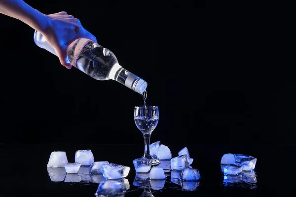 Woman pouring cold vodka from bottle into shot on black background