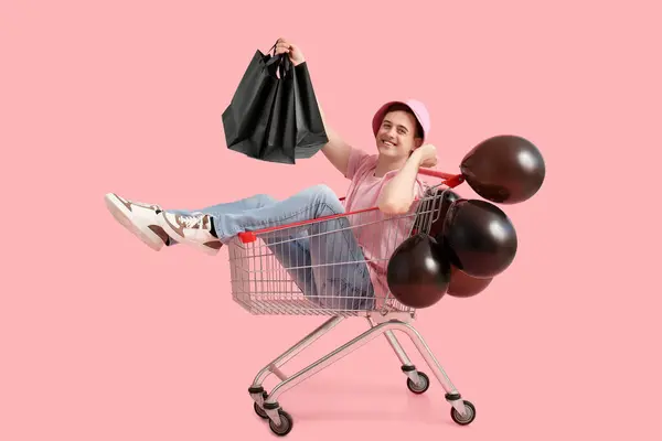 ピンクの背景にショッピングカートに座っているバッグと風船を持つ幸せな若者 ブラックフライデーセール — ストック写真