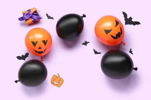 Different funny Halloween balloons with gift box and paper bats on lilac background