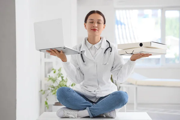 Female Asian doctor with laptop and folders meditating on table in clinic