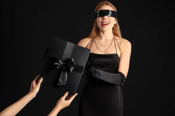 Young woman with blindfold receiving gift on dark background. Black Friday sale