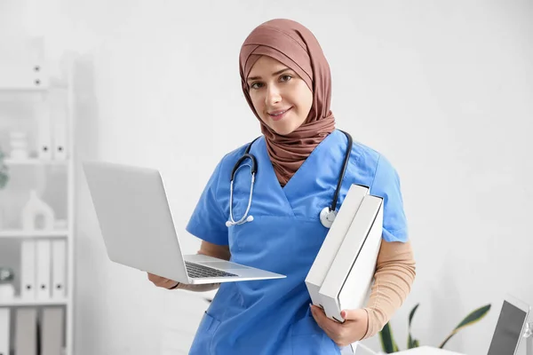 Female Muslim doctor with laptop and folders in clinic