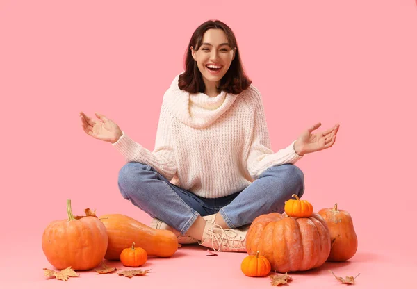 Young woman with pumpkins and autumn leaves sitting on pink background