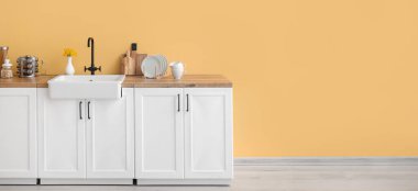 Sarı duvarın yanında lavabosu, sofra takımı ve ürünleri olan beyaz tezgahlar. Tasarım için pankart