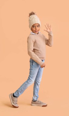 Sıcak kış kıyafetleri içinde, bej arka planda el sallayan sevimli Afro-Amerikalı çocuk.