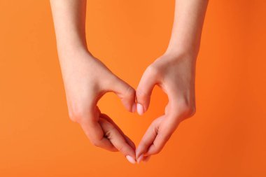Elleriyle turuncu arka planda kalp yapan bir kadın.