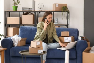 Evde dizüstü bilgisayarı olan genç bir kadın cep telefonuyla konuşuyor.