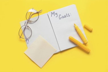 Listesi boş bir defter, sarı arka planda kalemler ve gözlükler. Yeni yıl hedefleri