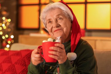 Noel Baba şapkalı yaşlı kadın Noel arifesinde evde kakao içiyor.