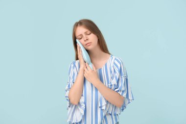 Dişi ağrıyan genç bir kadın mavi arka planda soğuk kompres tutuyor.