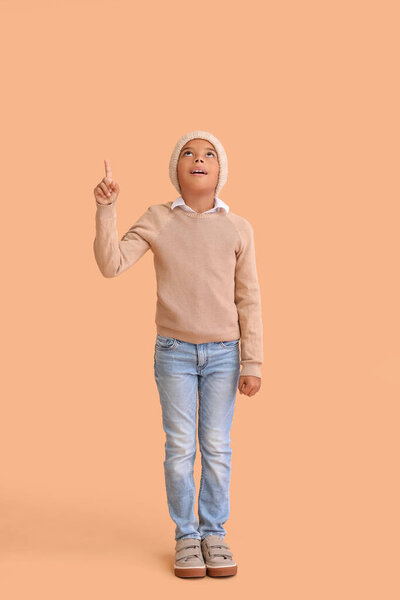 Cute African-American boy in warm winter clothes pointing at something on beige background