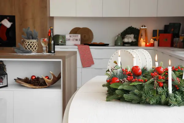 Christmas wreath with burning candles on white table in kitchen