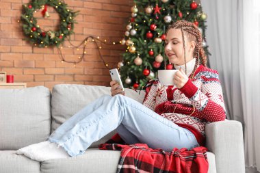 Noel arifesinde evde cep telefonu kullanan bir fincan kakaolu genç kadın.