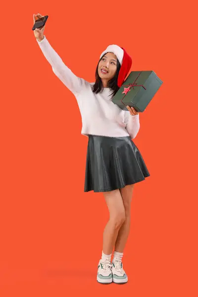 サンタの帽子の若い美しいアジアの女性は赤い背景のギフトボックスでセルフィーを取ります — ストック写真